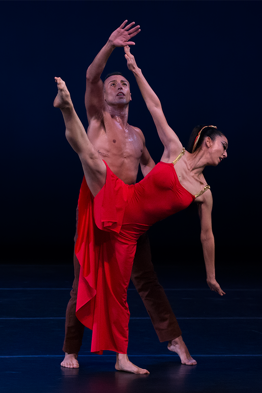 The woman in red pitches her body to the left side, kicking her right leg on a high right diagonal. Behind her her lover reaches upward almost touching her extend hand
